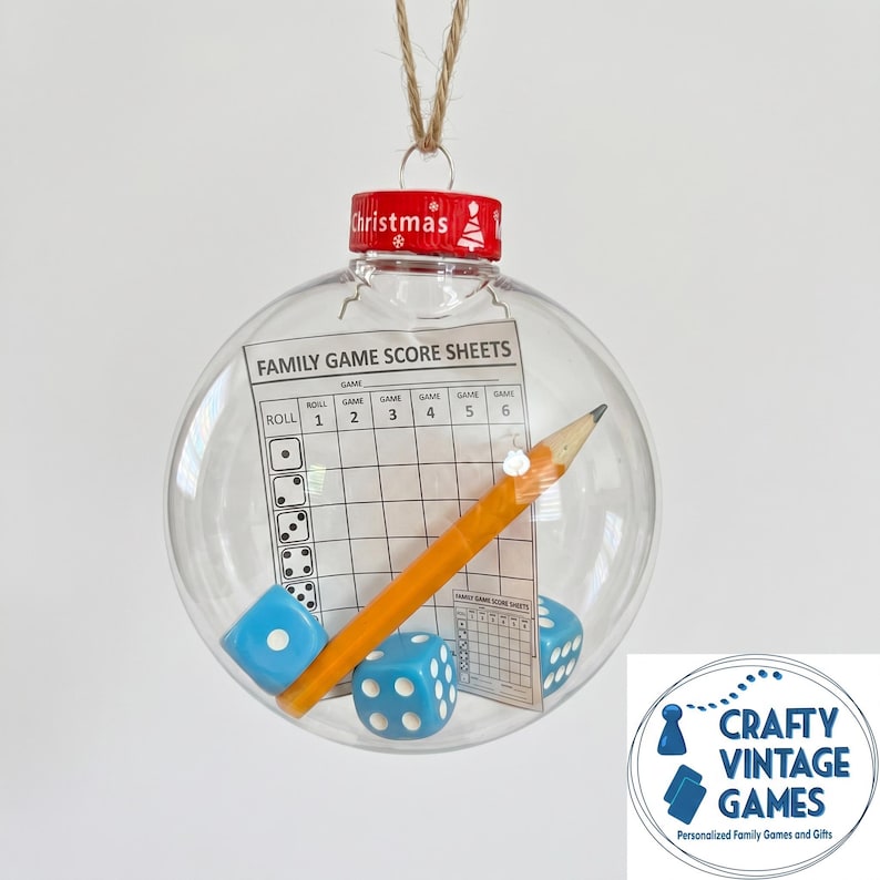 May include: A clear spherical Christmas ornament containing a printed family game score sheet, a yellow wooden pencil, and three light blue dice with white pips. The ornament has a red cap with the word Christmas and a small tree icon printed on it.