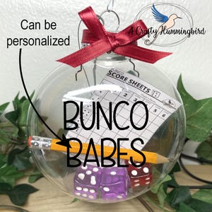 May include: A clear glass spherical ornament topped with a red satin ribbon bow. Inside the ornament are two purple dice, one red die, a yellow pencil, and a white Bunco score sheet. The text "BUNCO BABES" is printed on the front of the glass.