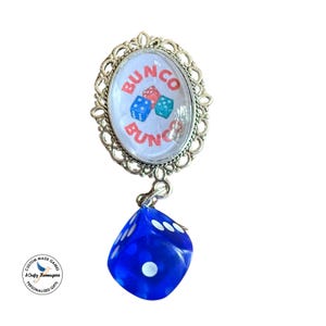 May include: An oval brooch with a silver-toned filigree frame featuring a white centre insert printed with the word BUNCO in red and three small dice. A single translucent blue die hangs from the bottom of the brooch as a dangling charm.