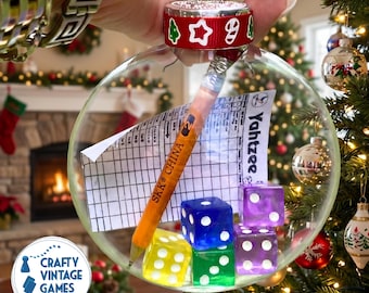 May include: A clear, spherical Christmas ornament filled with a Yahtzee score sheet, a yellow pencil marked SKK CHINA, and four translucent dice in yellow, blue, green, and purple. The ornament has a red ribbon with green and white icons.