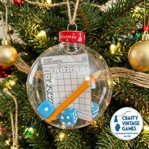 May include: A clear, spherical Christmas ornament containing a Bunco score sheet, a yellow pencil, and two light blue dice. The ornament features a red cap with the word Christmas printed in white text and hangs from a thin twine loop.