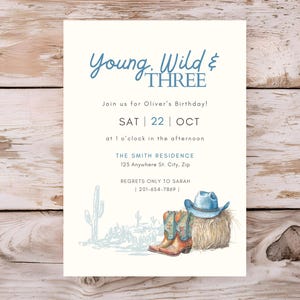 Könnte beinhalten: Eine Einladung zum Geburtstag mit dem Text "Young, Wild & THREE" in blauer Schrift. Die Einladung enthält Details für Olivers Geburtstag, mit einer Aquarellillustration von Cowboy-Stiefeln, einem Hut und einem Heuballen. Ein Kaktus ist ebenfalls abgebildet.