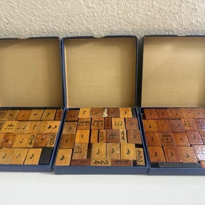 May include: Three open blue boxes containing a collection of rectangular wooden stamps. Each stamp features black ink illustrations of various musical notes, rests, and time signature numbers printed on the light brown wood surface.