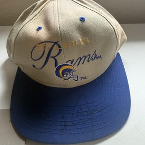 May include: A beige baseball cap with a blue brim and a blue button on top. The front features the embroidered text St. Louis Rams in blue and gold, accompanied by a small graphic of a ram helmet in blue, gold, and white with a TM symbol.