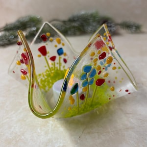 May include: A clear glass candle holder with a floral design. The holder features red, yellow, and blue flowers on a green background. The edges are outlined in green, and a white candle sits inside.