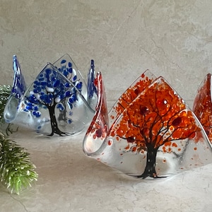 Può includere: Due portacandele in vetro con disegni di alberi. Uno presenta un albero blu, l'altro un albero arancione. I portacandele hanno una base trasparente e una forma triangolare ondulata. Gli alberi sono dipinti con tronchi e rami neri.