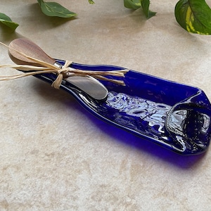 Könnte beinhalten: Eine blaue Glasflasche in Form einer Servierplatte mit einem Holzbuttermesser, das mit Schnur daran befestigt ist.