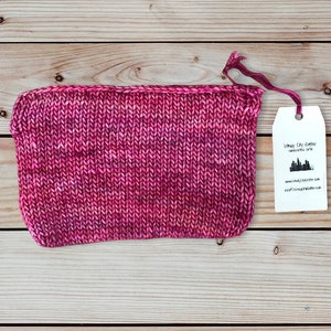 May include: A hand-knitted, rectangular pouch in shades of pink and purple, with a matching ribbon attached. A white tag with black text and a skyline graphic hangs from the ribbon. The pouch is made of soft yarn.