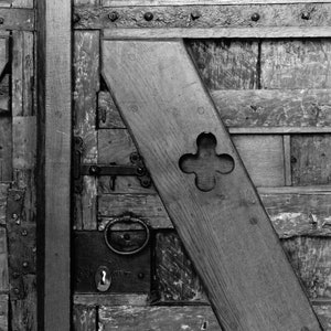May include: A black and white photograph of a wooden door with a metal latch and a decorative metal ring. The door is made of several different pieces of wood, and the wood is weathered and worn. The door has a metal strap across the top and a metal hinge on the left side.