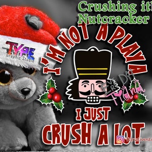 Pode incluir: Um gráfico de Natal vermelho e branco com uma ilustração de quebra-nozes e o texto "I'm not a playa I just crush a lot". O gráfico está sobre um fundo preto com um brinquedo de pelúcia cinza e branco no canto.