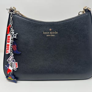 May include: Black Kate Spade shoulder bag with a silver chain strap. The bag features a charm with a Red Bull logo, a cowboy boot, and a Texas flag. The bag has the Kate Spade New York logo in gold.
