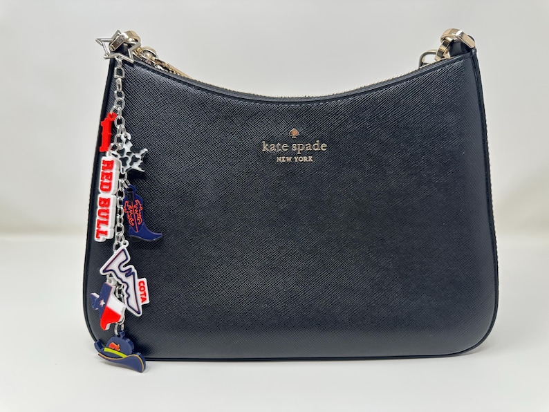 May include: Black Kate Spade shoulder bag with a silver chain strap. The bag features a charm with a Red Bull logo, a cowboy boot, and a Texas flag. The bag has the Kate Spade New York logo in gold.