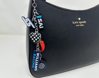 May include: Black leather handbag with a silver chain and multiple charms. The charms include a checkered heart, a red chili pepper, a blue and white "Williams Racing" tag, a blue and white "SAI" tag, and a black and white "55" tag. The bag has a gold "kate spade new york" logo.