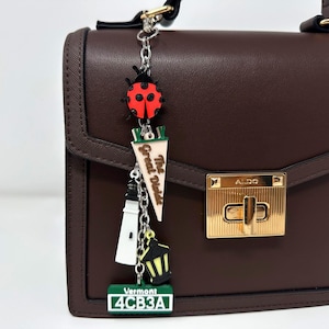 May include: A dark brown structured handbag with a gold-tone metal clasp labeled ALDO. A silver chain charm hangs from the handle, featuring a red ladybug, a pennant reading The Great Diode, a white lighthouse, a yellow lantern, and a Vermont license plate.