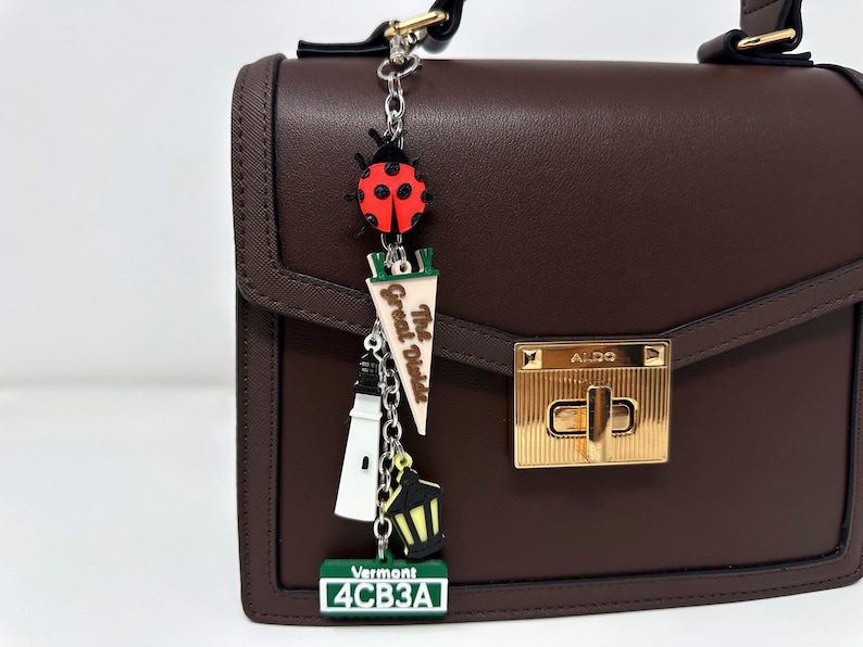 May include: A dark brown structured handbag with a gold-tone metal clasp labeled ALDO. A silver chain charm hangs from the handle, featuring a red ladybug, a pennant reading The Great Diode, a white lighthouse, a yellow lantern, and a Vermont license plate.