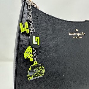 May include: Black leather handbag with a silver chain and a charm featuring a black and yellow helmet design, a yellow number four, and a black and white letter L. The bag has the text "kate spade new york" on the front.