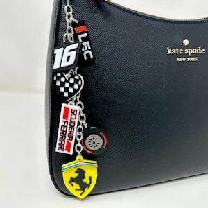 Könnte beinhalten: Schwarze Ledertasche mit goldenem 'kate spade new york' Logo. An der Henkeltasche hängt eine silberne Kette mit Anhängern, darunter ein rot-weißer '16' Anhänger, ein rot-weißer 'Scuderia Ferrari' Anhänger, ein schwarz-weiß kariertes Herz-Anhänger, ein gelber Ferrari-Logo-Anhänger und ein schwarz-silberner Reifen-Anhänger.