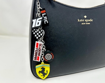 以下が含まれることがあります： 黒い革製のハンドバッグに、金色の「kate spade new york」のロゴが刻まれています。ハンドルには、銀色のチェーンにチャームがぶら下がっていて、赤と白の「16」のチャーム、赤と白の「スクーデリア・フェラーリ」のチャーム、黒と白の市松模様のハートのチャーム、黄色のフェラーリのロゴのチャーム、黒と銀のタイヤのチャームが付いています。