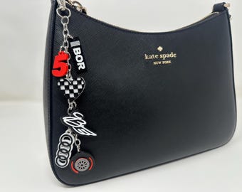 May include: Black Kate Spade shoulder bag with a silver chain and zipper. A decorative charm hangs from the chain, featuring a red number 5, a checkered heart, and other racing-themed charms. The bag has a gold logo.