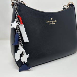 May include: Black handbag with a silver chain strap and a decorative charm. The charm features white stars, blue cowboy boots, and white and blue state shapes. The bag has the words "kate spade NEW YORK" in gold.