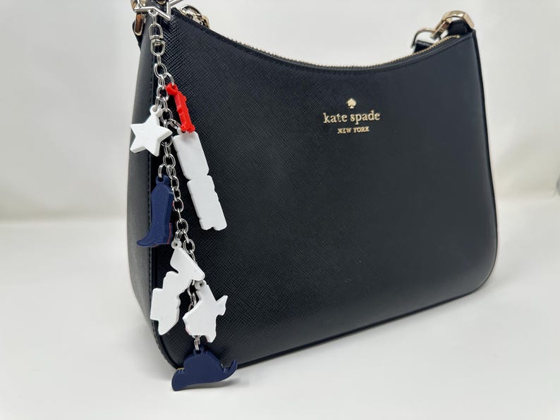 May include: Black handbag with a silver chain strap and a decorative charm. The charm features white stars, blue cowboy boots, and white and blue state shapes. The bag has the words "kate spade NEW YORK" in gold.