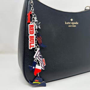 May include: A black Kate Spade handbag with a silver chain charm. The charm features a red and white "RED BULL" tag, a cowboy boot, a Texas flag, and a cowboy hat. The charm is attached to the handbag's zipper.