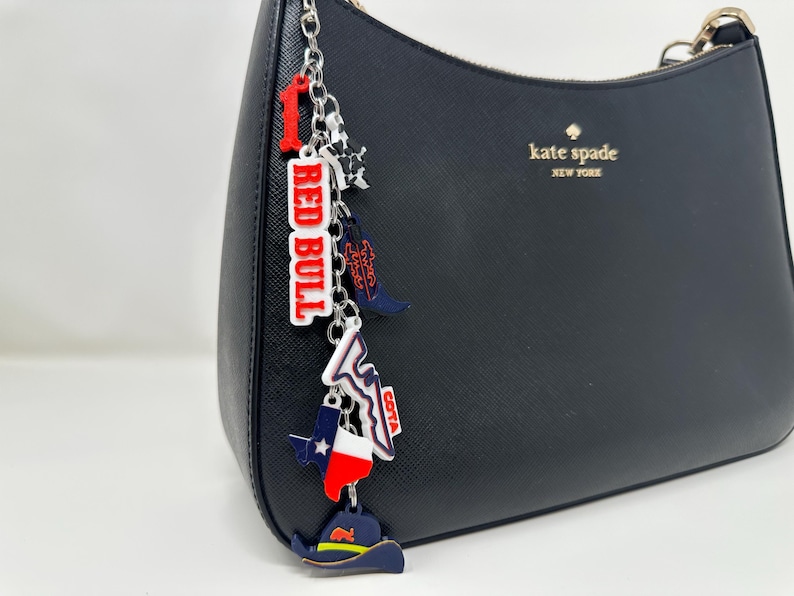 May include: A black Kate Spade handbag with a silver chain charm. The charm features a red and white "RED BULL" tag, a cowboy boot, a Texas flag, and a cowboy hat. The charm is attached to the handbag's zipper.