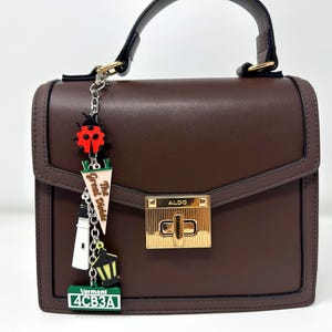 May include: A structured dark brown handbag with a gold-tone turn-lock closure labeled ALDO. A silver chain charm hangs from the handle, featuring a red ladybug, a pennant reading The Great Divide, a lighthouse, a lantern, and a Vermont license plate.