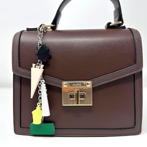 May include: A structured dark brown handbag with a top handle and a gold-tone metal turn-lock closure featuring the brand name ALDO. A silver-tone chain charm hangs from the side, displaying a black beetle, a beige triangle, a white tower, a yellow shape, and a green rectangle.
