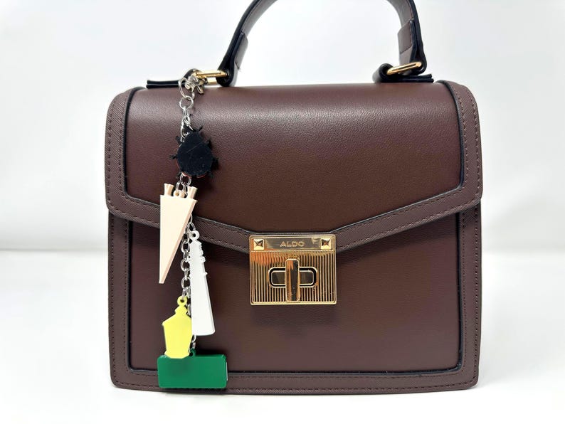 May include: A structured dark brown handbag with a top handle and a gold-tone metal turn-lock closure featuring the brand name ALDO. A silver-tone chain charm hangs from the side, displaying a black beetle, a beige triangle, a white tower, a yellow shape, and a green rectangle.