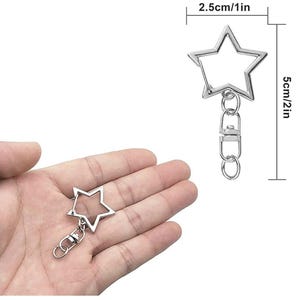 Pode incluir: Chaveiro prateado em forma de estrela com fecho mosquet&atilde;o e argola. A estrela mede 2,5 cm de largura e 5 cm de comprimento. O chaveiro &eacute; mostrado na m&atilde;o de uma pessoa.