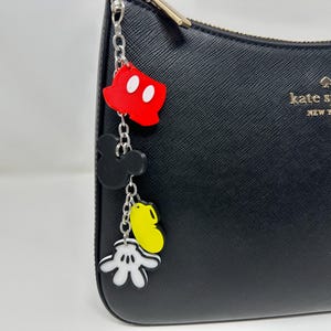 Puede incluir: Bolso de mano de cuero negro con una cadena plateada y un charm de Mickey Mouse. El charm representa la cabeza de Mickey Mouse, sus manos y un guante rojo. El bolso tiene el texto "kate spade new york" en la parte delantera.