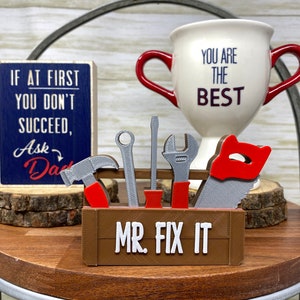 May include: A brown wooden toolbox with the text "MR. FIX IT" and miniature tools, including a hammer, wrench, screwdriver, and saw. A white mug with red handles and the text "YOU ARE THE BEST" is also present, along with a wooden sign that reads "IF AT FIRST YOU DON'T SUCCEED, Ask Dad."