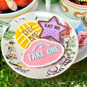 Puede incluir: Un plato decorativo con ilustraciones de Alicia en el país de las maravillas, con un círculo amarillo con el texto "TRY ME", una galleta en forma de estrella con "EAT ME" y una galleta rosa con "TAKE ONE". También se ve una pequeña botella con una etiqueta "DRINK ME".