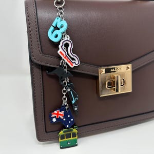 May include: A brown handbag with a gold clasp and a decorative keychain. The keychain features charms of a kangaroo, a tram, the Australian flag, and a race track. The handbag has a structured design.