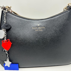 Pode incluir: Bolsa de ombro preta Kate Spade New York com z&iacute;per dourado e um berloque. O berloque inclui um l&aacute;bio preto, um c&iacute;rculo prateado, um cora&ccedil;&atilde;o vermelho, um c&iacute;rculo preto, uma estrela prateada e um ret&acirc;ngulo azul.