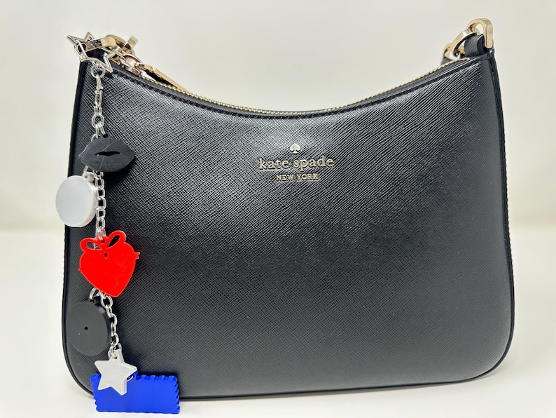 Pode incluir: Bolsa de ombro preta Kate Spade New York com z&iacute;per dourado e um berloque. O berloque inclui um l&aacute;bio preto, um c&iacute;rculo prateado, um cora&ccedil;&atilde;o vermelho, um c&iacute;rculo preto, uma estrela prateada e um ret&acirc;ngulo azul.