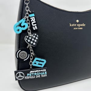 Könnte beinhalten: Schwarze Ledertasche mit einer silbernen Kette und Anhängern. Die Anhänger umfassen eine blaue Nummer 63, ein schwarz-weiß kariertes Herz, einen blauen Reifen, eine blaue Nummer 63 und einen blau-weißen Anhänger mit der Aufschrift "AMG PETRONAS FORMULA ONE TEAM". Die Handtasche hat ein goldenes "kate spade NEW YORK" Logo.