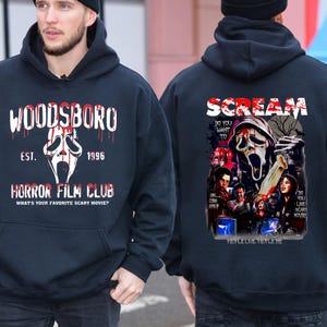 May include: A navy blue hoodie featuring the text "WOODSBORO" and "HORROR FILM CLUB" with a ghost face graphic. The back of the hoodie displays "SCREAM" with a movie poster design. The hoodie has a drawstring hood and a kangaroo pocket.