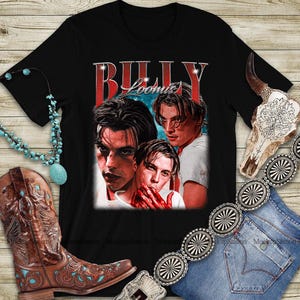 Puede incluir: Camiseta negra con un gr&aacute;fico vintage de tres hombres y el texto "BILLY LOOMIS" en rojo. La imagen se muestra con un collar turquesa, una bota de vaquero marr&oacute;n y un cintur&oacute;n plateado.
