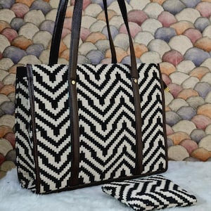 Op de afbeelding: Een zwarte en witte chevron patroon tote bag met bruine leren handvatten en afwerking. Een bijpassend klein etui staat voor de tas. De tas staat op een wit oppervlak met een kleurrijke achtergrond.
