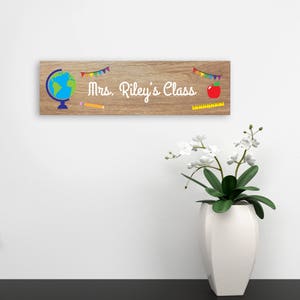 Puede incluir: Un letrero de madera con el texto "Mrs. Riley's Class" en letras blancas. El letrero est&aacute; decorado con un globo terr&aacute;queo, un l&aacute;piz, una regla, una manzana y una hilera de banderines coloridos.