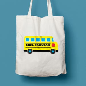 Puede incluir: Una bolsa de tela blanca con un dise&ntilde;o de autob&uacute;s escolar amarillo. El autob&uacute;s tiene ventanas azules y una se&ntilde;al de stop roja. El texto "MRS. JOHNSON" est&aacute; impreso en el lateral del autob&uacute;s.
