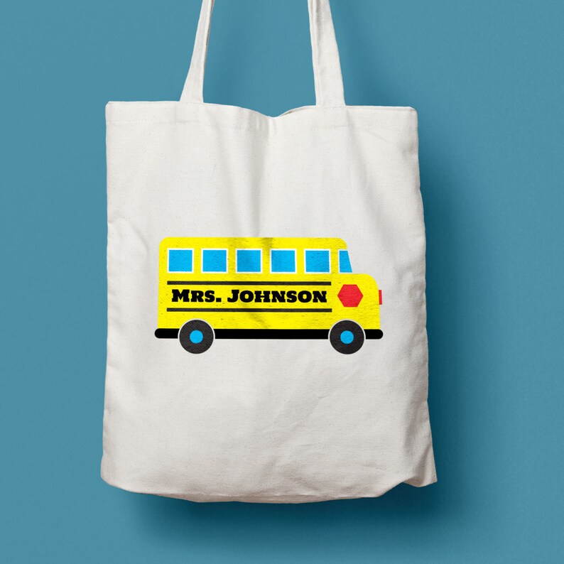 Puede incluir: Una bolsa de tela blanca con un dise&ntilde;o de autob&uacute;s escolar amarillo. El autob&uacute;s tiene ventanas azules y una se&ntilde;al de stop roja. El texto "MRS. JOHNSON" est&aacute; impreso en el lateral del autob&uacute;s.