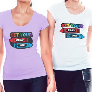 Puede incluir: Dos mujeres con camisetas que llevan el texto "Get Your Cray On!" impreso. Las camisetas tienen un dise&ntilde;o de crayones de colores.