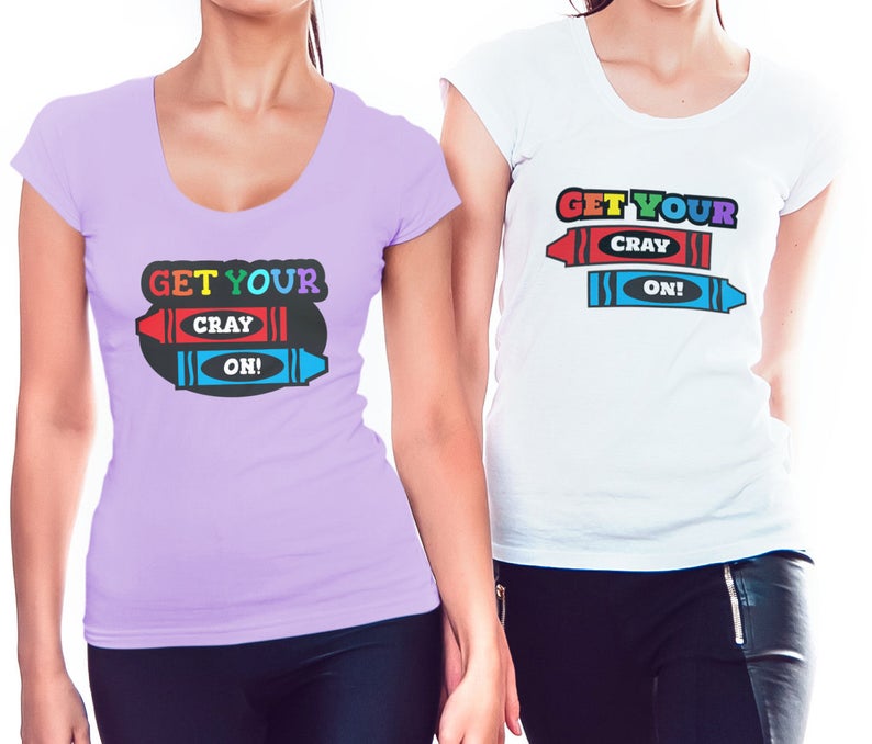Puede incluir: Dos mujeres con camisetas que llevan el texto "Get Your Cray On!" impreso. Las camisetas tienen un dise&ntilde;o de crayones de colores.