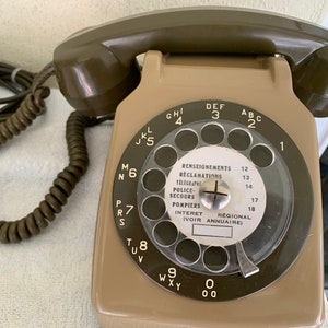 Puede incluir: Un teléfono de disco rotatorio verde oliva y beige con un cable en espiral. El dial tiene números y letras, con texto en francés que indica servicios de emergencia. Un dispositivo de comunicación vintage.
