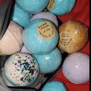 May include: A collection of handmade bath bombs in various colors, including blue, gold, lavender, and white, some with glitter and labels that read "Fruit loops handmade with love". The bath bombs are wrapped in clear plastic.