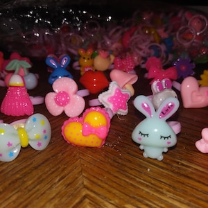 May include: A collection of colorful plastic rings with various designs, including hearts, stars, flowers, animals, and bows. The rings are all pink, blue, yellow, and white.