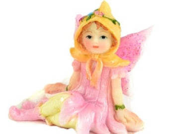 May include: A whimsical figurine of a fairy in a pink flower dress and matching wings. The fairy has a yellow bonnet and a yellow scarf. The fairy is sitting down. A charming decorative item.